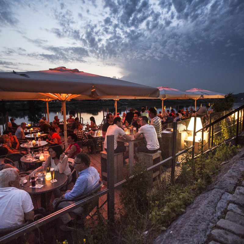 Groepen mensen zitten 's avonds op het terras aan de Maas met knusse verlichting en kaarsjes