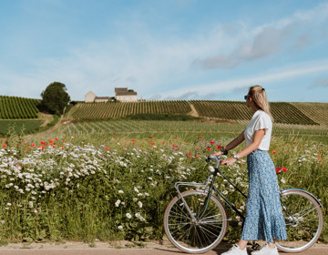 Een vrouw met lange rok en fiets wandelt over de weg en kijkt uit over een heuvellandschap met bloemen, wijngaarden en oude boerderij
